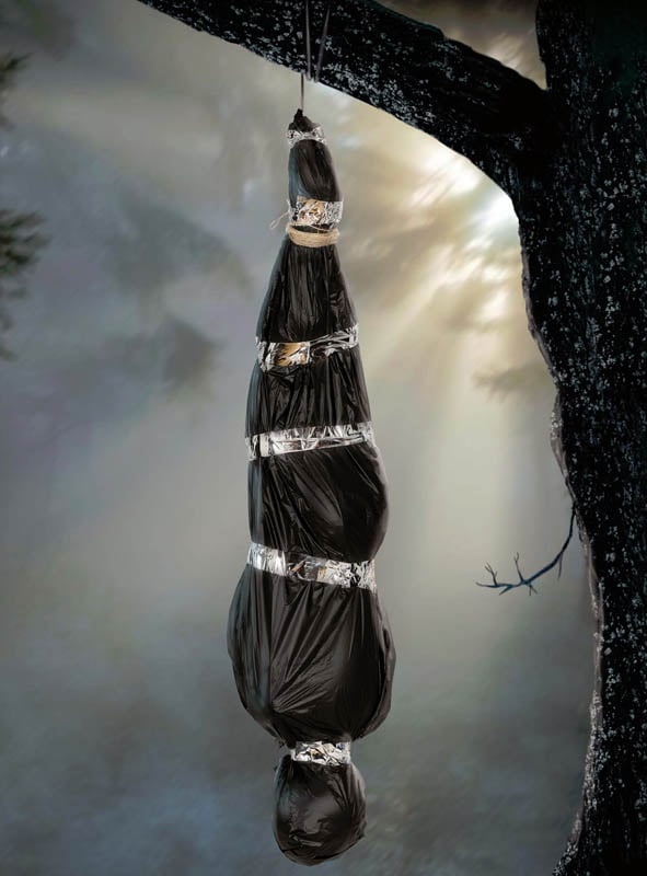 Eine beunruhigende „Aufblasbare Leiche im Leichensack 178 cm“ hängt kopfüber an einem Ast in einer schummrig beleuchteten Waldszene – ein schwarzer, aufblasbarer Leichensack in Menschengestalt, der sich perfekt als gruselige Halloween-Dekoration eignet.