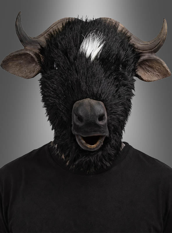 Eine Person, die die „Büffel Stier Vollmaske“ trägt – eine äußerst realistische schwarze Stiermaske mit markanten Hörnern und einem charakteristischen weißen Fleck –, steht in einem schlichten schwarzen Hemd vor einem grauen Hintergrund mit Farbverlauf. Diese auffällige „Büffel Stier Vollmaske“ eignet sich ideal für Maskenbälle oder Fasching-Feiern.