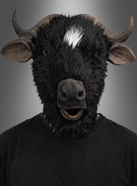 Eine Person, die die „Büffel Stier Vollmaske“ trägt – eine äußerst realistische schwarze Stiermaske mit markanten Hörnern und einem charakteristischen weißen Fleck –, steht in einem schlichten schwarzen Hemd vor einem grauen Hintergrund mit Farbverlauf. Diese auffällige „Büffel Stier Vollmaske“ eignet sich ideal für Maskenbälle oder Fasching-Feiern.