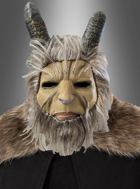 Vor einem grauen Farbverlaufshintergrund ist eine Person zu sehen, die die „Biest Vollmaske mit Hörner Krampus“ trägt, die mit ziegenartigen Hörnern, Fellverzierungen und einem braunen Umhang ausgestattet ist.