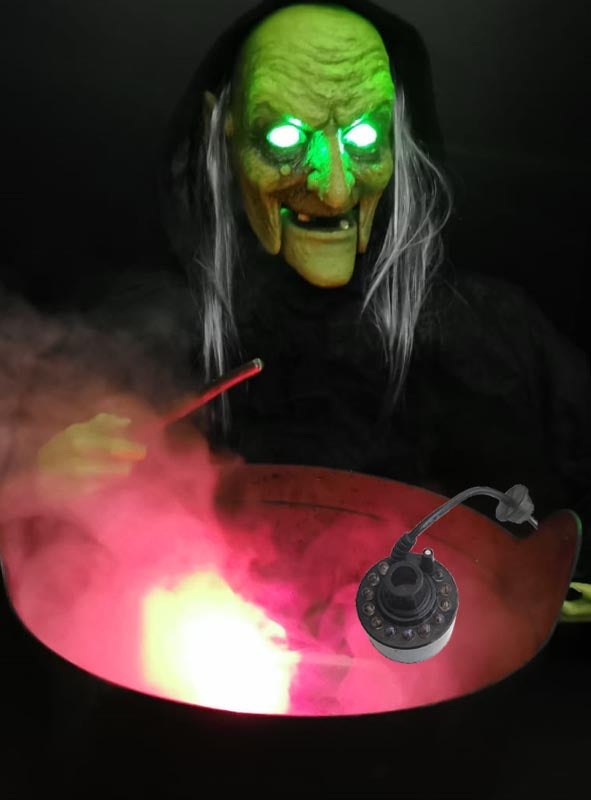 Eine Hexe mit grünem Gesicht und leuchtenden Augen steht mit einem Zauberstab in der Hand über einem rauchenden Kessel, aus dem rotes Licht strahlt; am Rand des Kessels ist eine kleine „Bunte LED Mini Nebelmaschine“ befestigt.