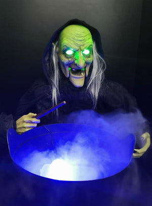 Eine Hexe mit grünem Gesicht, leuchtenden Augen und einem Zauberstab steht über einem Kessel, aus dem blaues Licht und Nebel strömen, wie bei einer „Bunte LED Mini Nebelmaschine“.