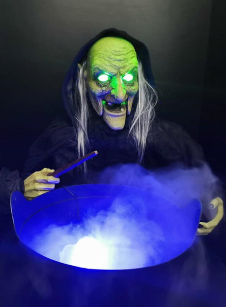 Eine Hexe mit grünem Gesicht, leuchtenden Augen und einem Zauberstab steht über einem Kessel, aus dem blaues Licht und Nebel strömen, wie bei einer „Bunte LED Mini Nebelmaschine“.