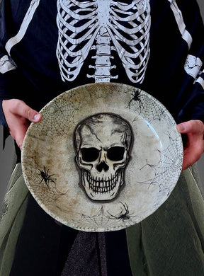 Eine Person in einem Skelettkostüm hält die XXL-Totenkopfschale (31 cm) für Halloween-Snacks in der Hand, die mit einer großen Totenkopf-Illustration, Spinnweben und Spinnen verziert ist – ideal zum Servieren von Halloween-Leckereien oder als gruselige Tischdekoration.