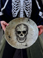 Eine Person in einem Skelettkostüm hält die XXL-Totenkopfschale (31 cm) für Halloween-Snacks in der Hand, die mit einer großen Totenkopf-Illustration, Spinnweben und Spinnen verziert ist – ideal zum Servieren von Halloween-Leckereien oder als gruselige Tischdekoration.