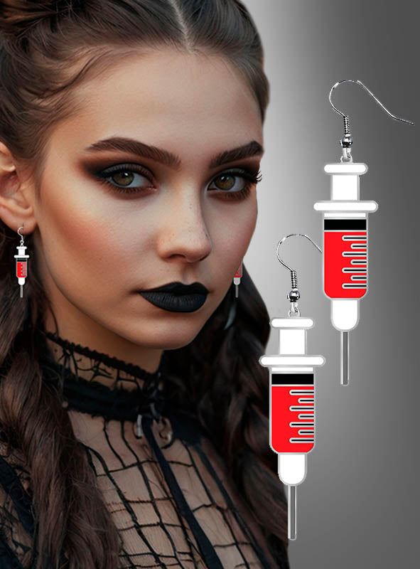 Eine Frau mit dunklem Make-up und welligem braunem Haar trägt die „Blutige Spritzen“-Ohrringe für Halloween – rot-weiße Ohrringe in Spritzenform, die sich perfekt als gruseliger Schmuck oder als Ergänzung zu einem Krankenschwester-Kostüm eignen – vor einem grauen Hintergrund mit Farbverlauf.