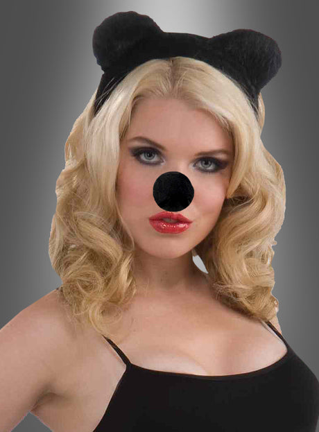 Eine Frau mit blonden Haaren, die ein schwarzes Stirnband mit Bärenohren und eine schwarze Maus-Schaumstoffnase trägt, gekleidet in ein schwarzes Oberteil, vor einem grauen Hintergrund mit Farbverlauf.