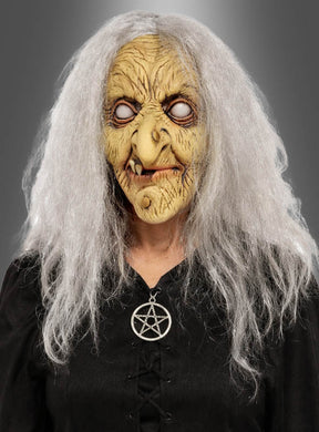 Sie trägt die „Hexen Maske Latex mit Haaren“ mit weißen Haaren, ist in schwarze Kleidung gekleidet und trägt eine Pentagramm-Halskette, vor einem grauen Hintergrund.