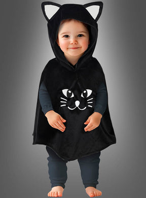 Ein Kleinkind, das einen „Süßes Kätzchen“-Poncho trägt – ein Babykostüm in Form eines schwarzen Katzenponchos mit einer Kapuze mit Katzenohren und einem Comic-Katzenkopf auf der Vorderseite –, steht barfuß vor einem grauen Hintergrund.
