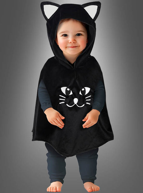 Ein Kleinkind, das einen „Süßes Kätzchen“-Poncho trägt – ein Babykostüm in Form eines schwarzen Katzenponchos mit einer Kapuze mit Katzenohren und einem Comic-Katzenkopf auf der Vorderseite –, steht barfuß vor einem grauen Hintergrund.
