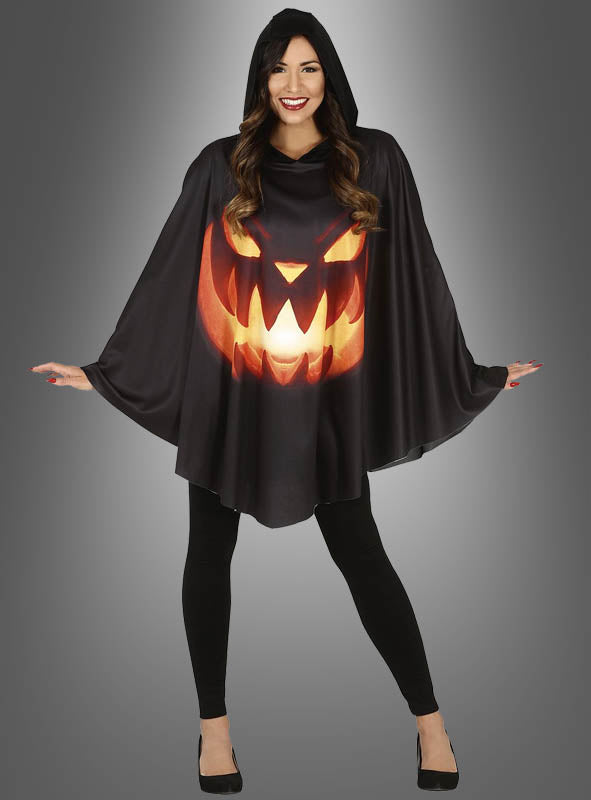 Eine Frau trägt das „Kürbis-Cape mit Kapuze Halloween“, einen schwarzen Poncho mit Kapuze, auf dessen Vorderseite ein leuchtendes Kürbiskopf-Gesicht abgebildet ist, dazu schwarze Leggings und schwarze Schuhe.