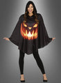 Eine Frau trägt das „Kürbis-Cape mit Kapuze Halloween“, einen schwarzen Poncho mit Kapuze, auf dessen Vorderseite ein leuchtendes Kürbiskopf-Gesicht abgebildet ist, dazu schwarze Leggings und schwarze Schuhe.