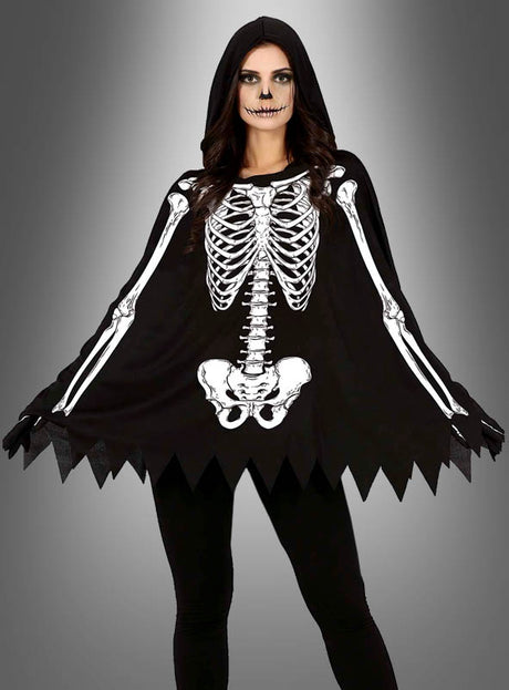 Eine Person im „Skelett-Poncho“-Halloween-Kostüm – bestehend aus einem schwarzen Poncho mit weißem Skelett-Aufdruck und Kapuze – steht mit einem als Totenkopf geschminkten Gesicht vor einem grau verlaufenden Hintergrund – eine ideale Wahl für Halloween-Kostüme für Damen bei jeder gruseligen Feier.