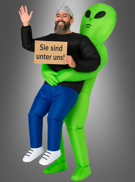 Eine Person mit einem Aluhut hält ein Schild mit der Aufschrift „Sie sind unter uns!“ in der Hand und trägt dabei ein aufblasbares Alien-Kostüm für Erwachsene, wodurch es so aussieht, als würde ein grüner Außerirdischer sie tragen.
