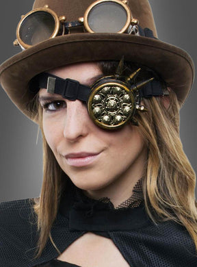 Eine schwarz gekleidete Person trägt einen braunen Steampunk-Hut mit Schutzbrille und einem verzierten goldenen Okular, elegant ergänzt durch das „Steampunk Monokel Unisex“ als stilvolles Steampunk-Accessoire, vor einem grauen Hintergrund mit Farbverlauf.