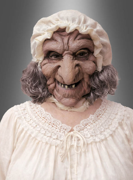 Eine Person, die die „Großmutter“-Latexmaske mit Haube trägt – mit übertriebenen Gesichtszügen, einer weißen Haube, grauen Haaren und einer weißen, mit Spitze besetzten Bluse –, posiert vor einem einfarbigen Hintergrund mit Farbverlauf.