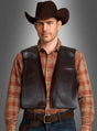 Ein Mann, der einen braunen Cowboyhut, ein braunes Karohemd, die braune Kunstleder-Cowboyweste und Jeans trägt, steht mit dem Gesicht nach vorne vor einem schlichten grauen Hintergrund.