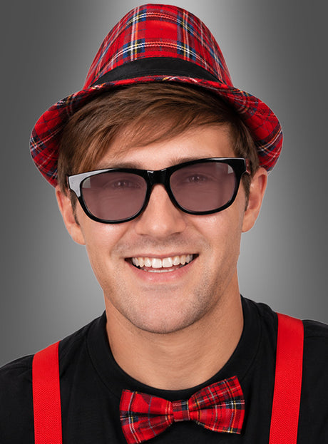 Eine lächelnde Person mit schwarzem Hemd, roter Karamütze, passender roter Karofliege, roten Hosenträgern und der getönten „Brille 50er 60er Jahre“ – im klassischen Nerdbrillen-Stil – vor einem grauen Hintergrund mit Farbverlauf.