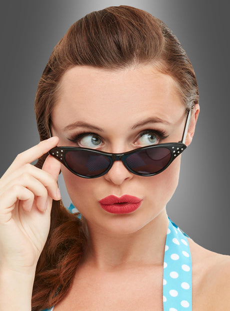 Eine Frau mit braunen Haaren und rotem Lippenstift, die die Sonnenbrille „50er Jahre schwarz“ und ein blaues Top mit Polka-Dots trägt und vor einem grauen Hintergrund nach oben und zur Seite blickt – ein perfektes Rockabilly-Accessoire, das den klassischen Stil der 50er Jahre verkörpert.