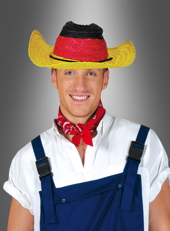 Ein Mann, der den Cowboy-Strohhut „Deutschland“ trägt – der mit seinem schwarz-rot-gelben Design optimalen Sonnenschutz bietet –, kombiniert mit einem roten Bandana, einem weißen Hemd und blauen Hosenträgern, lächelt vor einem grauen Hintergrund mit Farbverlauf.