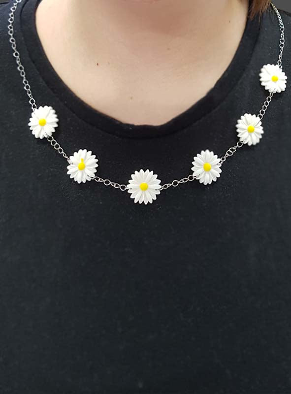 Eine Person, die ein schwarzes Shirt und die „Blumen-Kette Margerite“ trägt – eine Halskette mit sieben weißen und gelben Anhängern in Gänseblümchenform –, verkörpert perfekt die ausgelassene Stimmung einer Accessoire-Party oder einer lebhaften 70er-Jahre-Mottoparty.