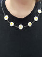 Eine Person, die ein schwarzes Shirt und die „Blumen-Kette Margerite“ trägt – eine Halskette mit sieben weißen und gelben Anhängern in Gänseblümchenform –, verkörpert perfekt die ausgelassene Stimmung einer Accessoire-Party oder einer lebhaften 70er-Jahre-Mottoparty.