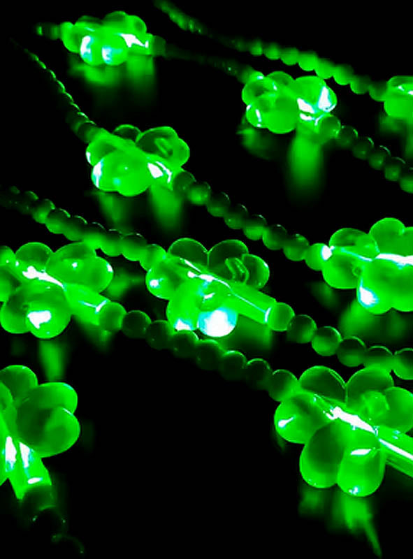 Die „Kleeblatt-Kette mit LED“ besteht aus grünen, kleeblattförmigen Lichtern an Perlenketten, die vor einem dunklen Hintergrund leuchten und in diagonalen Reihen angeordnet sind.
