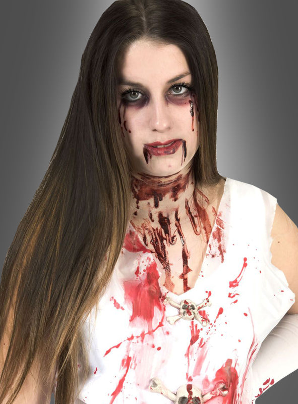Eine Frau mit langen Haaren in einem weißen Outfit, über und über mit Kunstblut bespritzt und mit dramatischem Make-up, zeigt Narben-Tattoos und realistische Bluteffekte im Gesicht und am Hals, die mit dem Wundenset mit Kunstblut erzeugt wurden, und posiert vor einem grauen Hintergrund.