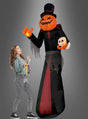 Eine Frau betrachtet den „Gigantischen Kürbis 365 cm, aufblasbar mit Licht“ – eine riesige aufblasbare Halloween-Figur mit Kürbiskopf, leuchtender Hand, zerfetzter schwarzer Robe und Zylinder, die einen kleinen Totenkopf in der Hand hält – und die im Garten eine auffällige Halloween-Dekoration bildet.