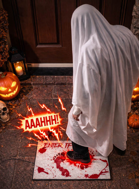 Eine Person in einem Geisterkostüm tritt auf die „Türmatte Soundeffekt für Halloween“ – eine blutbefleckte Fußmatte mit der Aufschrift „ENTER IF YOU DARE“ –, wodurch ein gruseliger roter „AAAHHH!“-Soundeffekt ausgelöst wird, während daneben Kürbisse und ein Totenkopf zu sehen sind.