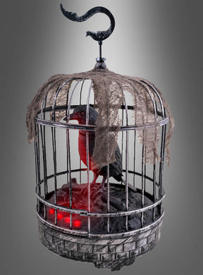 Ein schwarzer Vogel sitzt in einem Metallkäfig mit zerfetztem Stoff und rot leuchtenden Lichtern am Boden, vor einem grauen Hintergrund mit Farbverlauf – eine ideale Halloween-Dekoration mit dem „Sprechenden Raben im Käfig 32 cm (animiert)“, die jeder gruseligen Szene eine schaurige Atmosphäre verleiht.