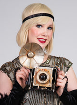 Eine Frau mit blondem Bob-Haarschnitt hält eine Retro-Dekokamera mit Blitz in der Hand und lächelt den Betrachter an. Sie trägt ein mit Perlen besticktes Flapper-Kleid und ein Stirnband im Stil der 1920er Jahre – ideal für eine Vintage-Dekoration oder eine unvergessliche 20er-Jahre-Party.