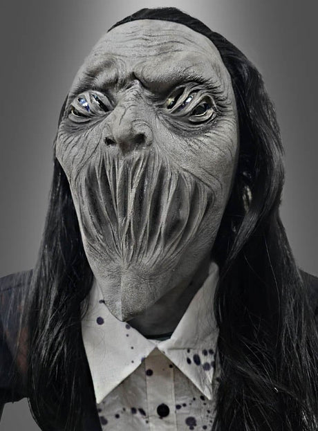 Die Figur trägt eine detailreiche „Gigantischer Zombie 230 cm“-Animatronik-Maske mit verzerrten Gesichtszügen, hohlen Augen und langen vertikalen Schlitzen als Mund und erinnert an einen hoch aufragenden Friedhofsgeist. Mit ihrem langen schwarzen Haar und einem Hemd mit Kragen vermittelt sie den gruseligen Eindruck einer beunruhigenden Halloween-Figur, die zum Leben erweckt wurde.