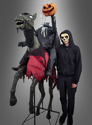 Eine Person mit einer Totenkopfmaske und einem schwarzen Kapuzenpulli steht neben der Halloween-Dekoration „Gigantischer kopfloser Reiter, 210 cm, Pferd, animiert“, die den kopflosen Reiter auf einem schwarzen Pferd zeigt, der einen Kürbiskopf in der Hand hält.