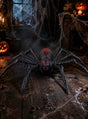 Eine große schwarze Spinne mit roten Flecken auf dem Rücken sitzt auf einem Holzboden in einem schummrigen Raum, der mit Spinnweben, Kerzen und geschnitzten Kürbissen geschmückt ist und so die perfekte Halloween-Dekoration bildet. Die „Terror Spinne 70 cm Tarantula mit Licht & Ton“ verleiht der gruseligen Atmosphäre eine lebendige, unheimliche Note.