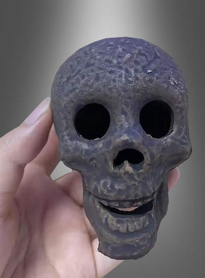 Eine Hand hält einen kleinen, dunklen, verwitterten, feuerfesten Keramik-Totenkopf mit großen Augenhöhlen und einem leicht geöffneten Mund vor einem grau verlaufenden Hintergrund – ein faszinierendes Halloween-Dekostück.