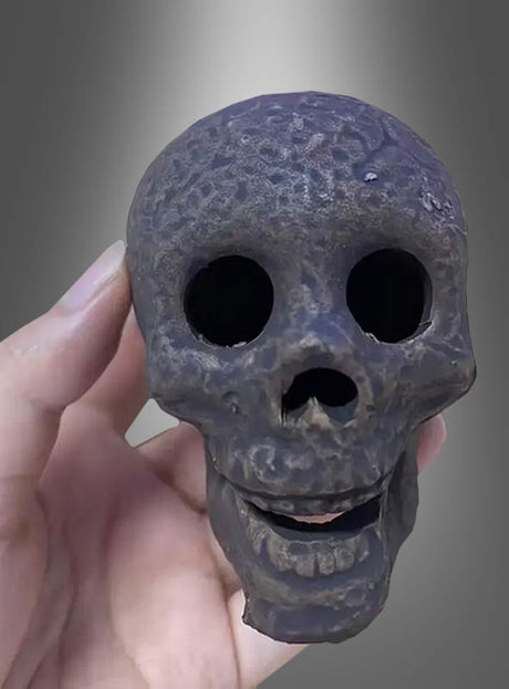 Eine Hand hält einen kleinen, dunklen, verwitterten, feuerfesten Keramik-Totenkopf mit großen Augenhöhlen und einem leicht geöffneten Mund vor einem grau verlaufenden Hintergrund – ein faszinierendes Halloween-Dekostück.