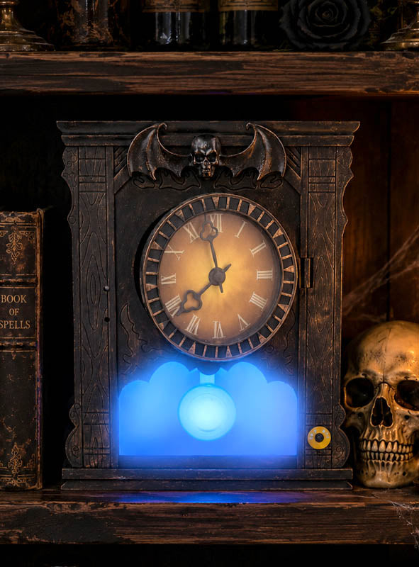 Auf einem Regal steht eine kunstvoll verzierte Uhr im gotischen Stil mit Fledermaus- und Totenkopfmotiv, deren Sockel blau leuchtet, neben einem Totenkopf und einem alten Buch mit der Aufschrift „SPELLS“ – eine perfekte „Animierte Geisteruhr 25 cm“ als Halloween-Dekoration für Ihre gruselige Dekoration.
