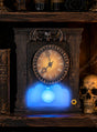 Auf einem Regal steht eine kunstvoll verzierte Uhr im gotischen Stil mit Fledermaus- und Totenkopfmotiv, deren Sockel blau leuchtet, neben einem Totenkopf und einem alten Buch mit der Aufschrift „SPELLS“ – eine perfekte „Animierte Geisteruhr 25 cm“ als Halloween-Dekoration für Ihre gruselige Dekoration.
