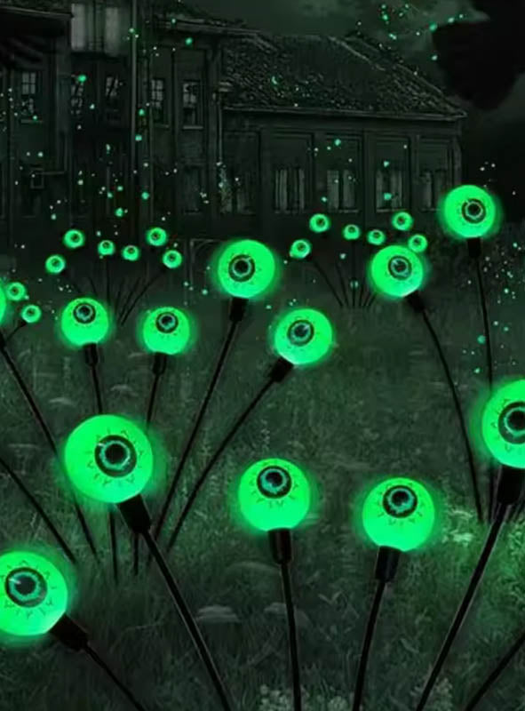 Vor einem dunklen, verlassenen Gebäude ist nachts ein Feld mit „Leuchtende Solar Augen“-Gartendekoration zu sehen – leuchtend grüne, augenähnliche Kugeln an Stielen –, die sich perfekt als gruselige Gartendekoration eignet.