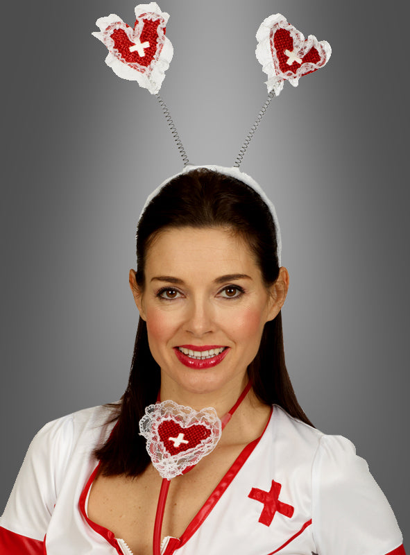 Eine Frau lächelt vor einem schlichten grauen Hintergrund. Sie trägt ein Krankenschwester-Kostüm mit einem herzförmigen Krankenschwester-Haarreif und einem passenden herzförmigen Fasching-Accessoire um den Hals.