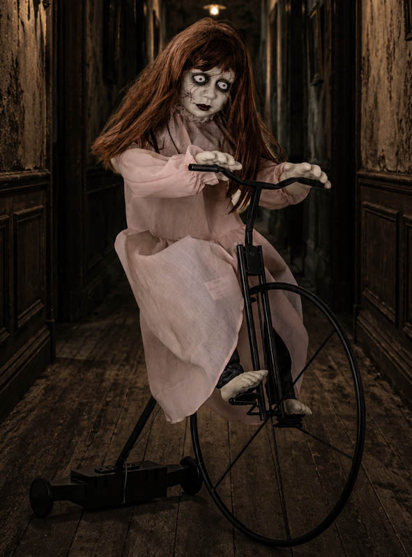 Die animierte Zombie-Puppe 72 x 94 cm auf einem Dreirad sitzt mit ihrem blassen Gesicht, den roten Haaren und dem rosa Kleid lebensecht auf einem altmodischen Dreirad in einem schmalen, düsteren Flur mit abgenutzten Holzböden und Wänden – eine unheimliche Halloween-Dekoration, die alle Blicke auf sich zieht.