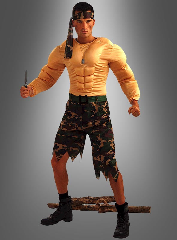 Ein Mann posiert im „Soldat“-Muskelkostüm im Rambo-Stil, komplett mit Muskeloberteil, Camouflage-Shorts, Stiefeln und einem Stirnband. Er hält ein Messer in der Hand und steht vor einem grauen Hintergrund mit Farbverlauf über einem Ast.
