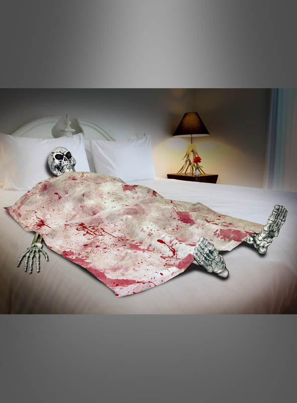Eine „Skelett im Bett“-Halloween-Dekoration liegt auf einem Bett, das mit einem blutigen Tuch bedeckt ist, in einem schwach beleuchteten Schlafzimmer, im Hintergrund sind eine Lampe und Kissen zu sehen.