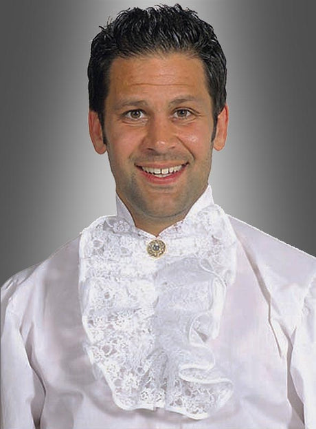 Vor einem grauen Hintergrund mit Farbverlauf steht ein Mann, der das Modell „Jabot aus Spitze New Romantic“ trägt – ein weißes Rüschenhemd mit Spitzendetails und einer dekorativen Brosche –, das dem Outfit einen zusätzlichen Hauch von Eleganz verleiht.