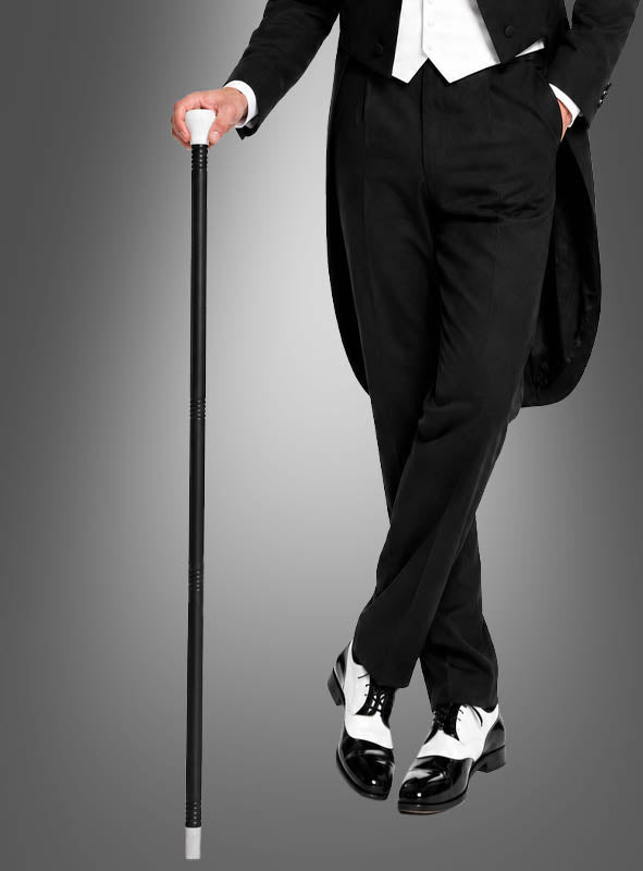 Ein vornehmer Herr in einem schwarzen Smoking mit weißen Handschuhen hält den „Tanzstock für Herren 100 cm extra lang“ in der Hand und steht mit übereinandergeschlagenen Beinen vor einem grauen Hintergrund – ein ideales Accessoire im Stil der 20er Jahre für jeden stilbewussten Mann.