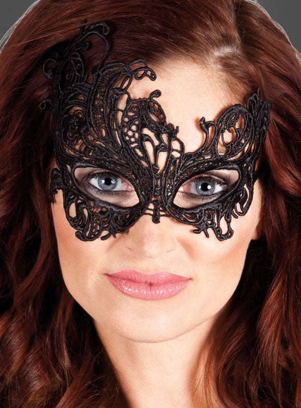 Eine Frau mit welligem rotem Haar trägt die Augenmaske „Fantasy“ aus schwarzer Spitze und blickt vor einem schlichten dunklen Hintergrund direkt in die Kamera.