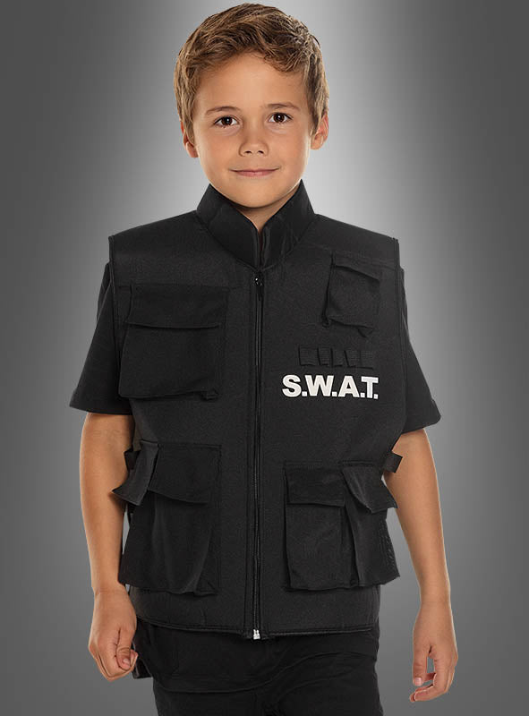 Ein kleiner Junge steht lächelnd vor einem einfarbigen grauen Hintergrund mit Farbverlauf und trägt dabei eine schwarze SWAT-Weste für Kinder.