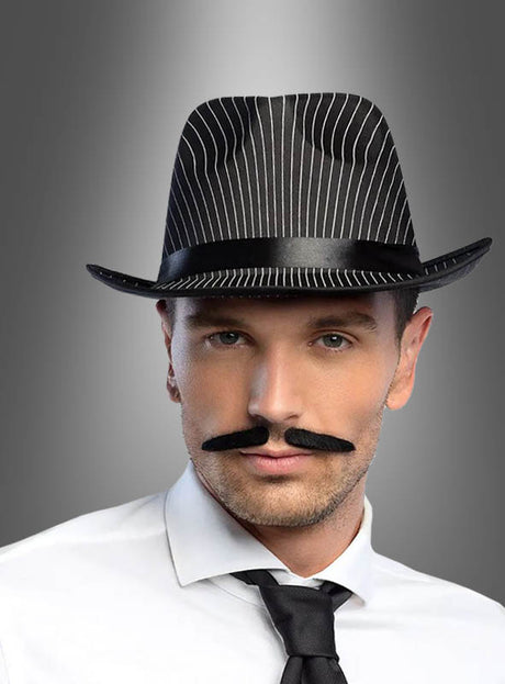 Ein Mann trägt den „Al Capone Mafia Bart“ – einen schwarzen, selbstklebenden Bart im klassischen Al-Capone-Stil –, dazu einen Nadelstreifen-Fedora, ein weißes Hemd und eine schwarze Krawatte, und verkörpert so den ikonischen Mafia-Look der 20er Jahre vor einem grauen Hintergrund mit Farbverlauf.