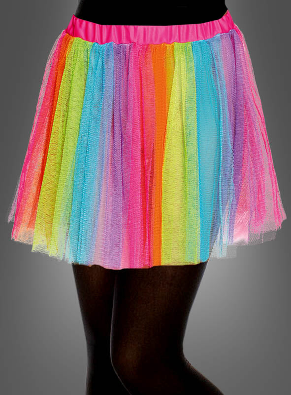 Ein Kind präsentiert den „Bunter Tüllrock Regenbogen Kinder“ – einen kurzen, regenbogenfarbenen Tüllrock mit vertikalen Streifen, der an ein Regenbogen-Tutu erinnert – kombiniert mit schwarzen Strumpfhosen vor einem grauen Hintergrund. Dieses farbenfrohe Ensemble ist perfekt für Fans von Einhorn-Kostümen für Mädchen oder alle, die auf der Suche nach einem Outfit mit einem bunten Tüllrock für Kinder sind.